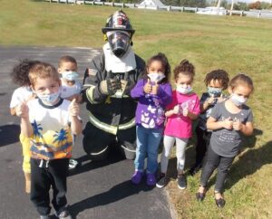 Preschool-aged children wearing surgical masks stand with a firefighter in full gear.  They are on the edge of a parking lot near a field.  The day is sunny.